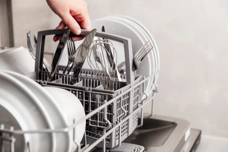 close-up of female hand holding full cutlery basket with clean knife, fork, whisk loading to, empty out or unloading from open automatic dishwasher machine with clean utensils in home kitchen