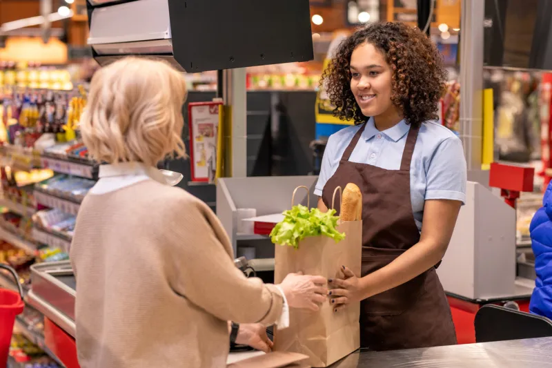 pretty young cashier giving mature female paperbag with bread and fresh groceries while both standing by cash register in supermarket