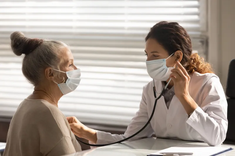 caring young female doctor in medical facemask hold stethoscope listen to elderly patient heart in hospital woman gp or physician do regular checkup examine mature client at consultation in clinic