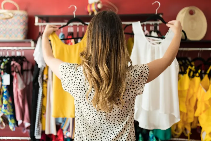 rear view of young female customer with long brown hair choosing between tops at shopping mall
