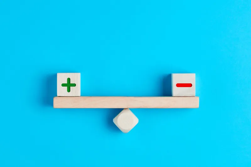 plus and minus or positive and negative symbols on wooden blocks are in balance on a wooden seesaw blue background, flat lay view pros and cons equilibrium in decision making under uncertainity