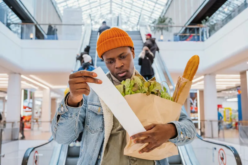 surprised african-american man in denim jacket looks at receipt total in sales check holding paper bag with products in mall