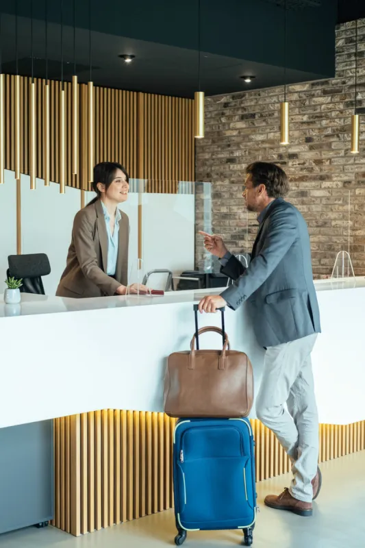business man with luggage talking with concierge on hotel reception with sneeze guard protection