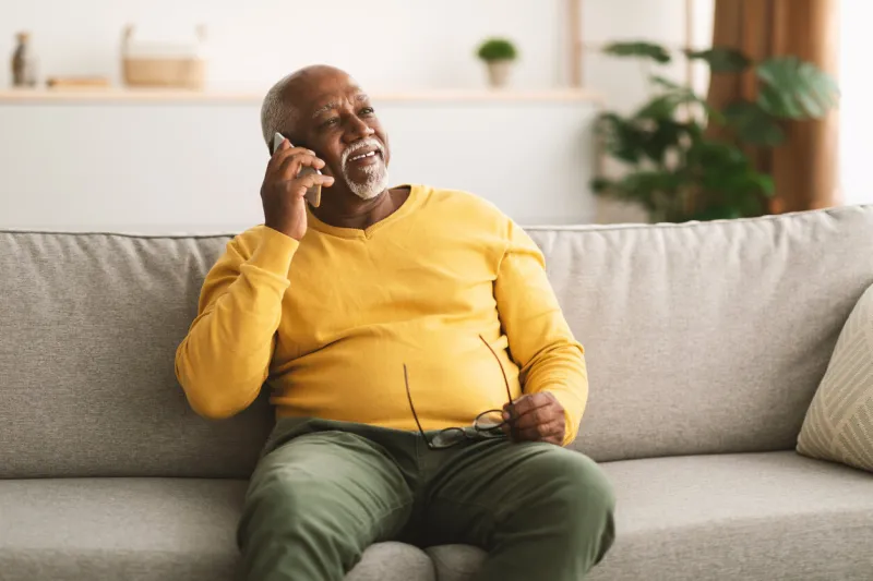 senior african american man talking on cellphone having pleasant phone conversation sitting on sofa at home, looking aside and smiling mobile communication concept