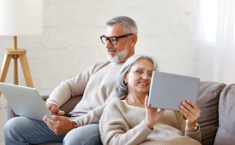 happy relaxed old retired family couple using modern technologies laptop and digital tablet while relaxing resting on sofa in living room, senior man and woman surfing internet in free time