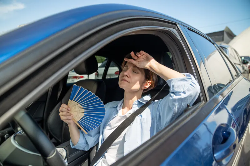 exhausted tired middle aged woman drives car waves blue fan suffers from stuffiness stands in urban traffic jam in summer hot weather overheating, high temperature in car with broken air conditioner