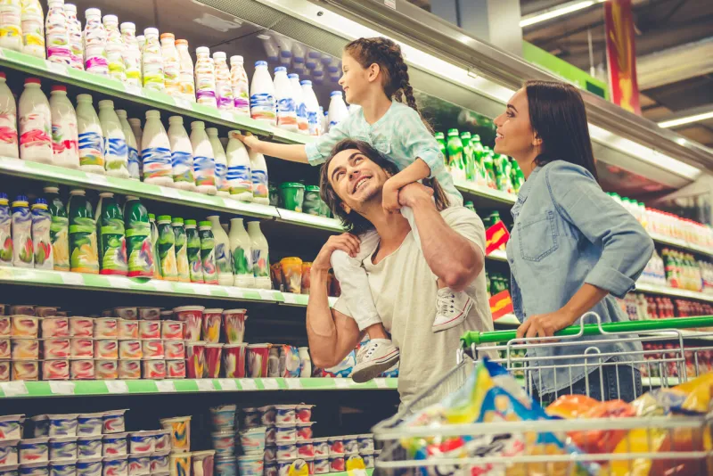 beautiful young parents and their cute little daughter are smiling while choosing food in the supermarket girl is sitting on her dad's shoulders