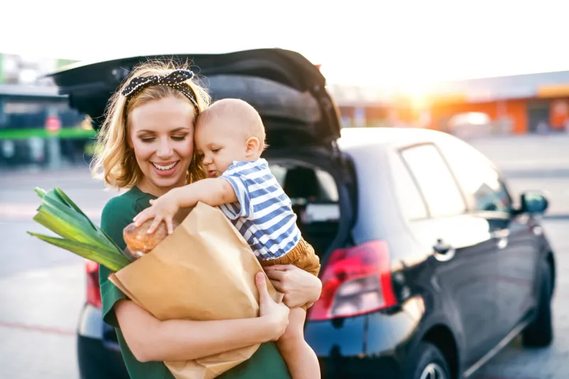beautiful young mother with her little baby son in front of a supermarket, holding paper shopping bag woman with a boy standing by the car