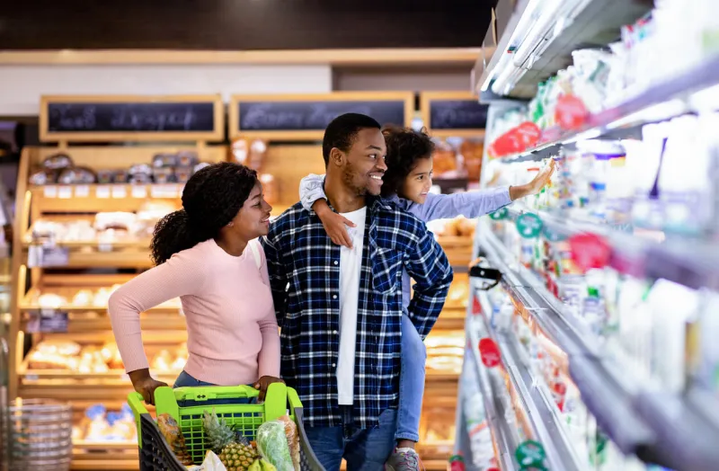 portrait of happy black family with trolley shopping together at grocery store millennial african american parents with lovely daughter picking food, choosing milk products at big mall