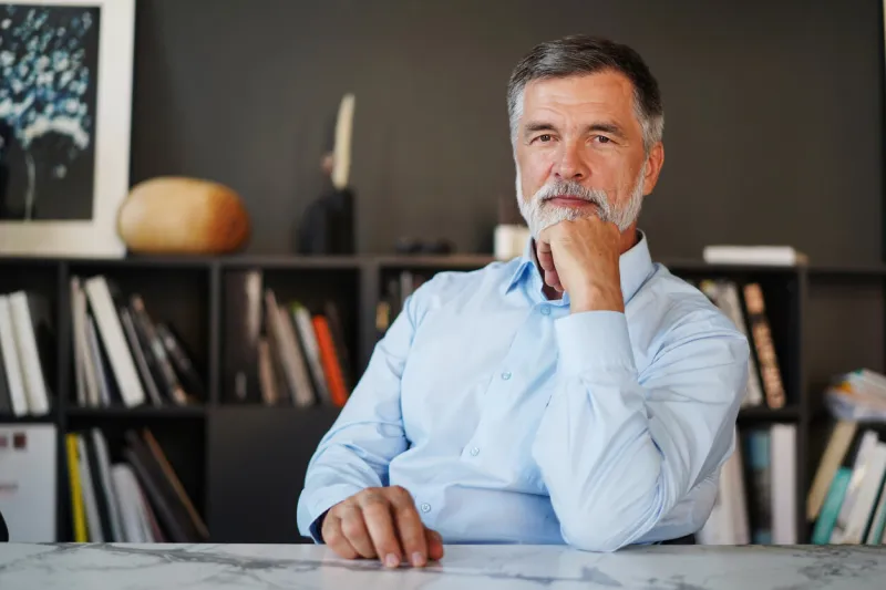 portrait mature professional businessman with grey hair looking at camera and smiling confident adult entrepreneur, leader, manager sitting in office