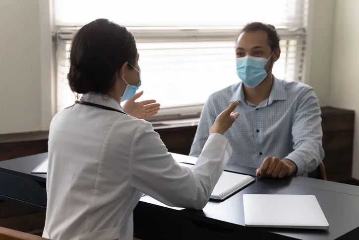 focused young african american male patient in protective facemask listening to skilled female indian general practitioner doc physician, discussing disease treatment or health test results in clinic