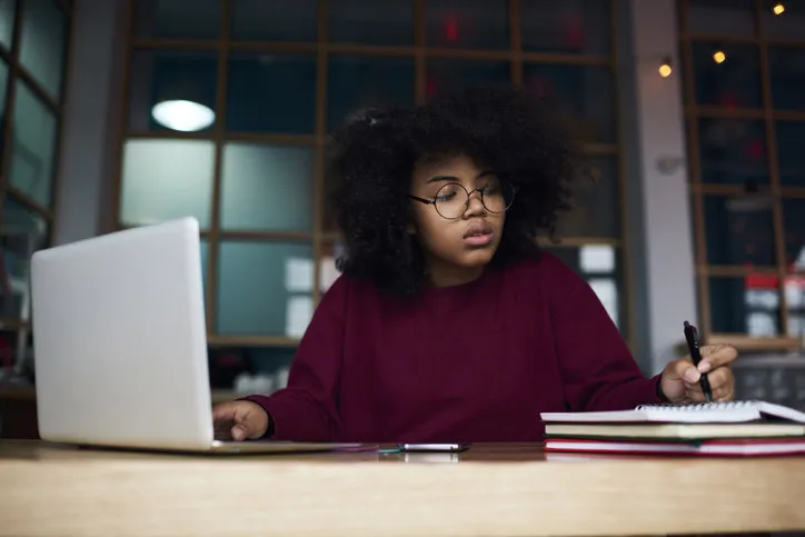 concentrated young female student in glasses learning in university campus using laptop computer and wireless connection to internet preparing to examination, attractive hipster girl writing essay