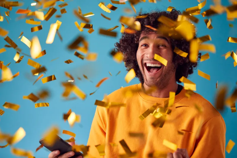 celebrating young man with mobile phone winning prize and showered with gold confetti in studio