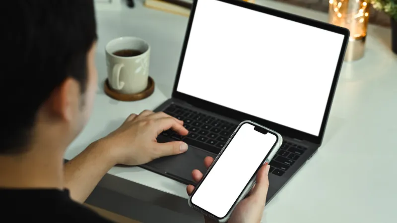 view over shoulder of man freelancer holding smart phone and using laptop at workplace empty display for advertising