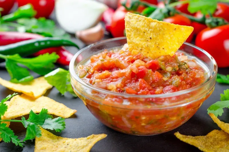 traditional mexican homemade salsa sauce with ingredients, tomatoes, pepper, cilantro, chips on a dark black stone table close-up, horizontal image