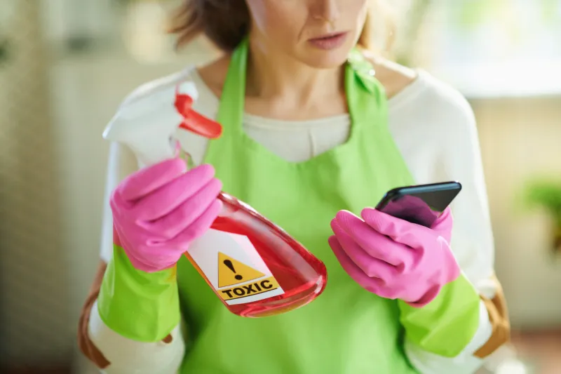 closeup on woman in green apron and pink rubber gloves in the living room in sunny day reading about cleaning product on internet using smartphone