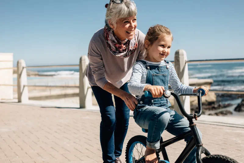 girl trying to ride a bicycle with the help of her grandmother at seaside promenade senior woman holding the bicycle while her granddaughter learns to ride it