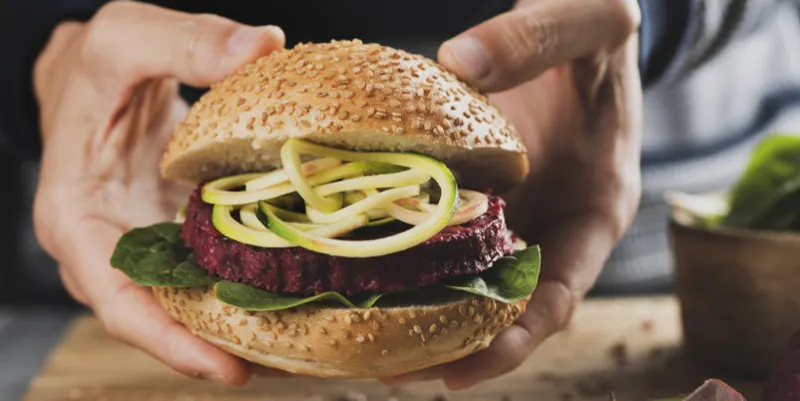 closeup of a young caucasian man with a beet burger sandwich in his hands, next to a rustic wooden table and a sliced beet