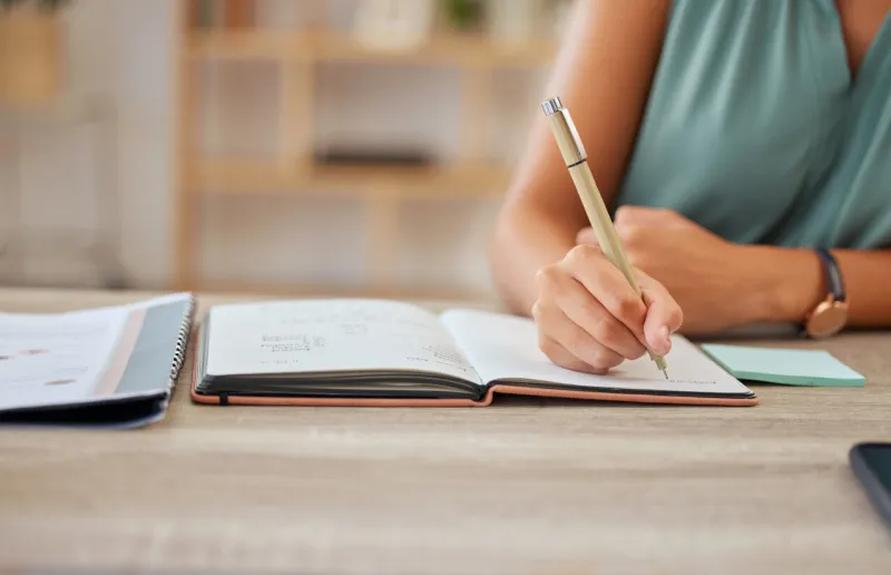 hand, pen and writing in a notebook with a business woman sitting at a desk in her office for planning agenda, schedule and appointment with a female employee making a note in her journal or diary