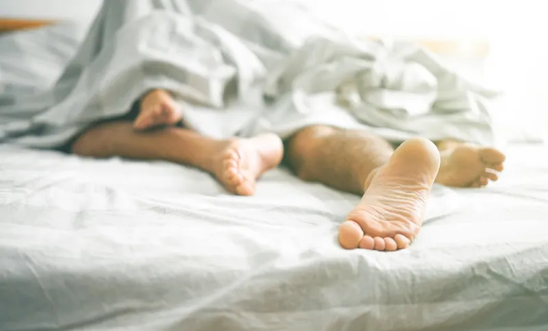 close up of male and female feet on a bed - loving couple having sex under under white blanket in the bedroom - concept of sensual and intimate moment of lovers - vintage filter - focus on male foot