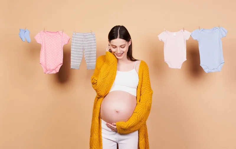 a nice, smiling young girl with long dark hair, in a yellow knitted cardigan, with a bare belly, touches her belly with her hands and looks at him with a sweet smile motherhood, childhood concept