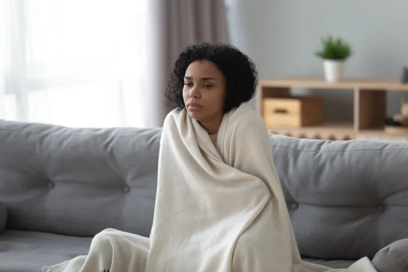 sick young african woman feeling cold covered with blanket sit on sofa, ill black girl shivering freezing warming at home wrapped with plaid, no central heating problem, fever temperature flu concept