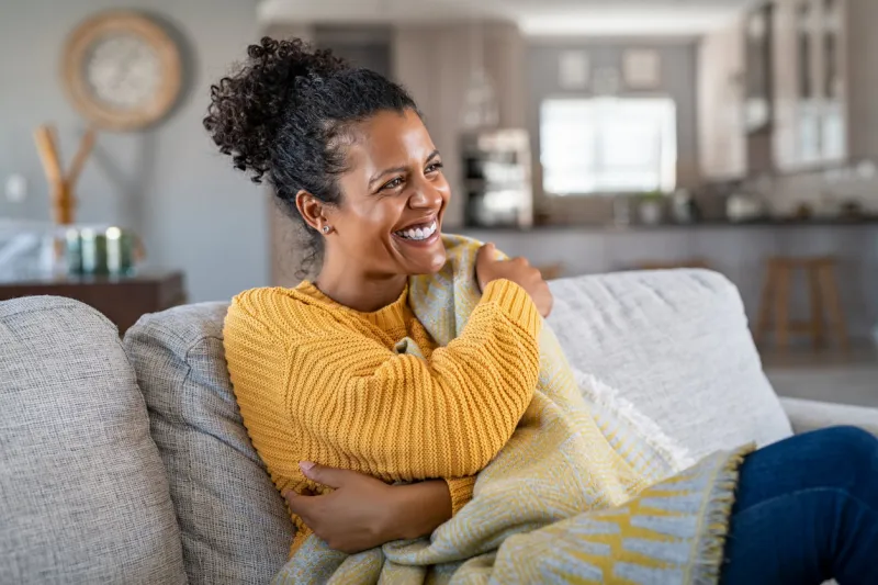 beautiful black girl sitting on couch wrapped under blanket and laughing cheerful african american woman sensitive to the cold relaxing at home on sofa carefree and happy mid woman hugging self with warm blanket in winter