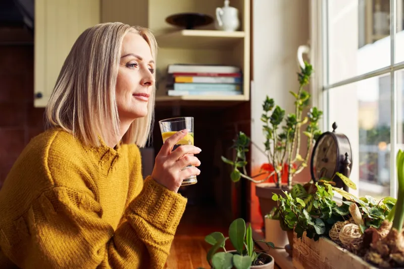mature woman drinking glass of water in the kitchen looking through the window