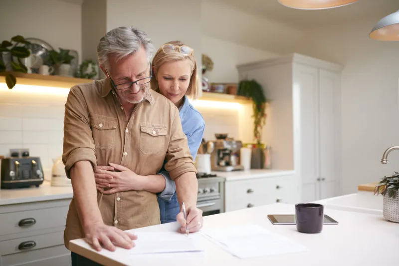 mature couple reviewing and signing domestic finances and investment paperwork in kitchen at home