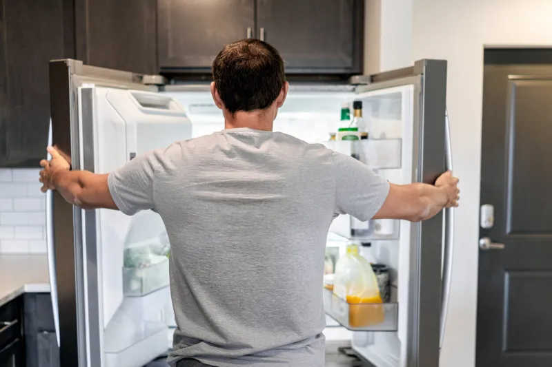 back of hungry man opening fridge refrigerator doors domestic appliance searching for food inside with condiments and juice in modern kitchen