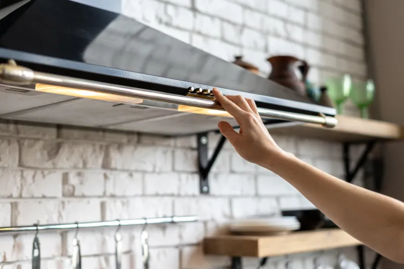 cropped view of woman hand select mode on cooking hood, standing near kitchen appliance in contemporary interior with brick wall and decor on shelves at blurred background