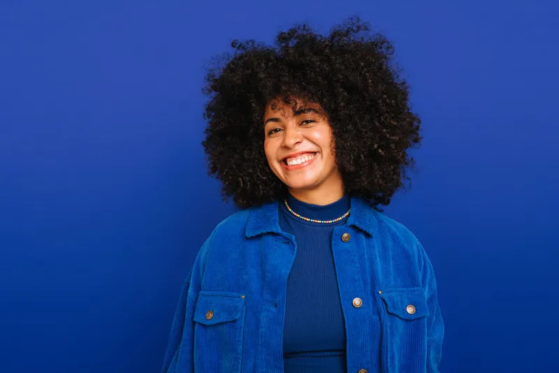 feeling good in blue happy woman with curly hair smiling at the camera while standing against a blue background beautiful woman with curly hair wearing her natural air with pride