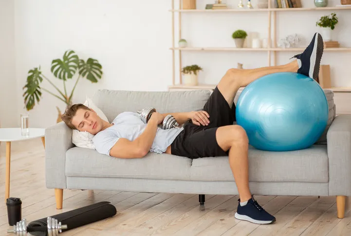 laziness and workout tired guy sleeping on couch with dumbbell and fitness ball, in living room interior