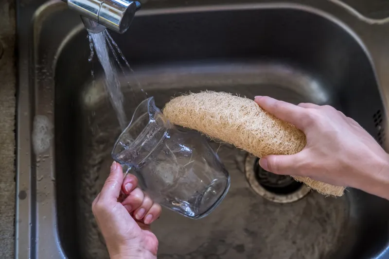 woman hands cleaning glass cup with natural compostable loofah sponge by the sink with running tap water eco-friendly kitchen and home products zero waste and sustainable plastic free lifestyle