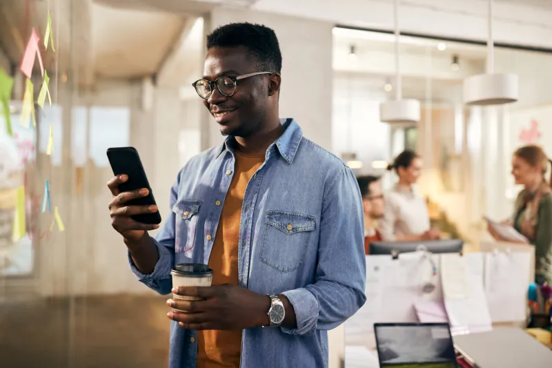 happy black entrepreneur using smart phone while having coffee break in the office his colleagues are in the background