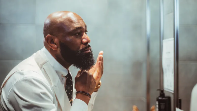 a profile view of a handsome bald black man grooming, brushing, and moisturizing the beard hair in front of the mirror in a bathroom of a luxury hotel, an african-american man taking care of his beard