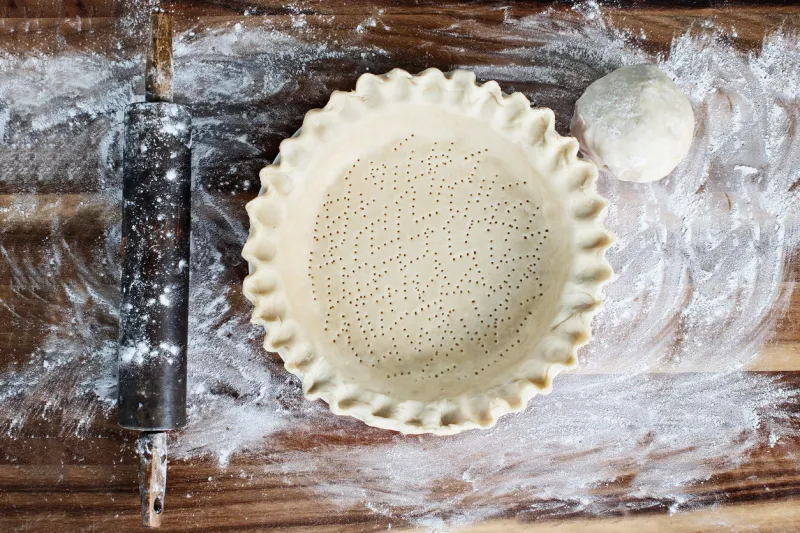 homemade butter pie crust in pie plate with fluted pinched edge, rolling pin and extra ball of dough over floured rustic wooden background crust has been perforated with fork and ready for baking