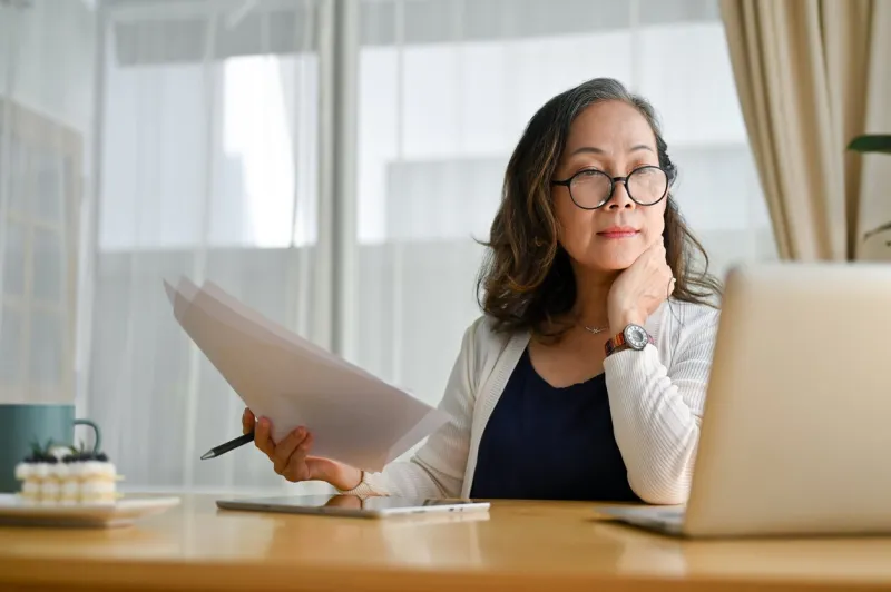 concentrated asian middle aged female teacher or businesswoman in glasses sitting at desk using portable computer and examining paperwork age and technology