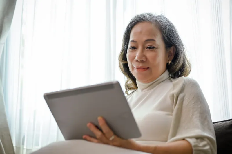 a beautiful asian-aged woman watches a video clip online on a digital tablet touchpad in her comfortable living room