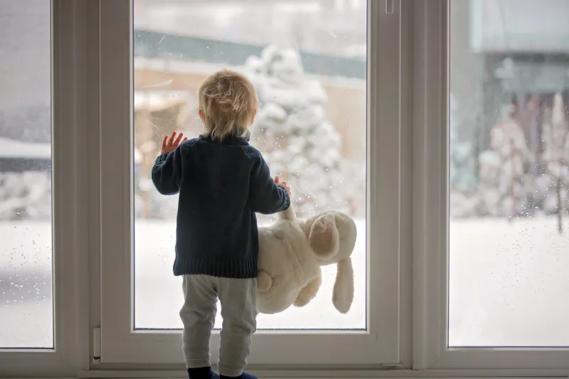 toddler child standing in front of a big french doors, leaning against it looking outside at a snowy nature, holding teddy toy