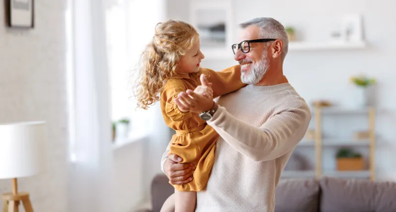 elegant loving caring grandfather looking at his cute little granddaughter, adorable child girl and positive grandpa holding hands while dancing together in living room at home family concept