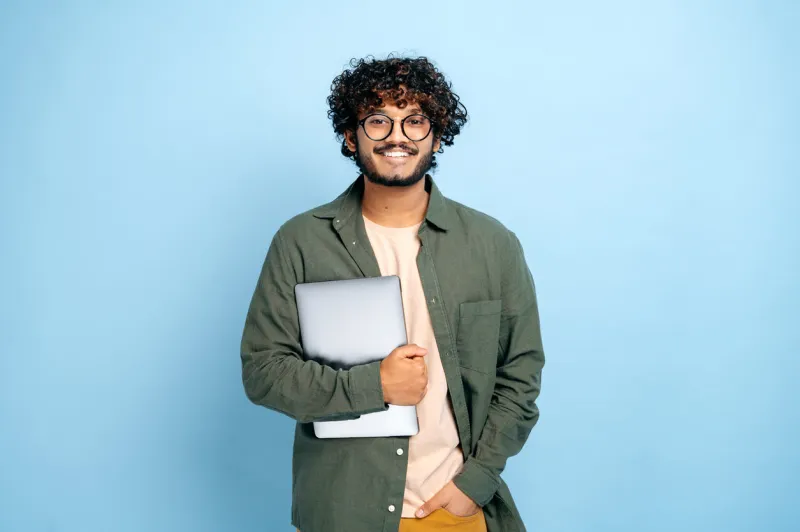 smart handsome positive indian or arabian guy, with glasses, in casual wear, student or freelancer, holding a laptop in hand, standing on isolated blue background, looking at camera, smiling friendly
