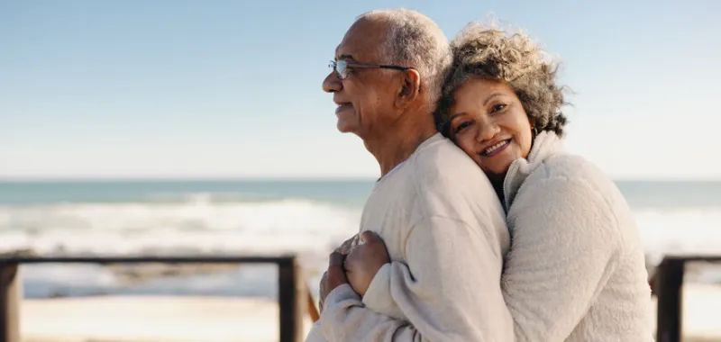 romantic senior woman smiling at the camera while embracing her husband by the ocean affectionate elderly couple enjoying spending some quality time together after retirement