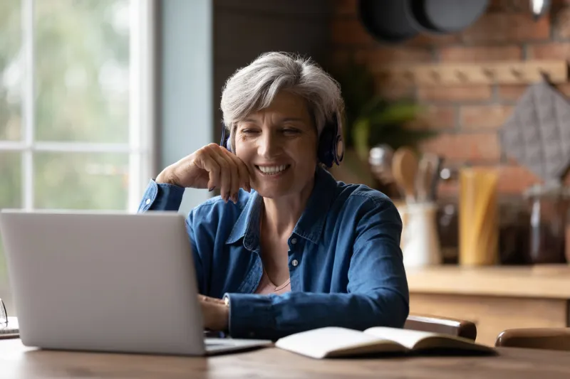smiling modern mature 60s caucasian woman in earphones look at laptop screen watch webinar online at home happy senior grey-haired female have fun study on internet elderly technology concept