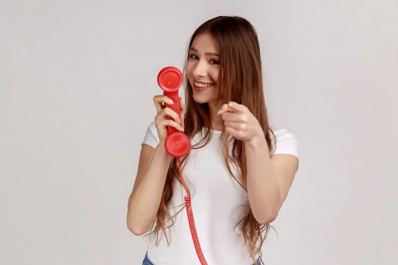 smiling attractive woman holding handset, showing call me gesture looking at camera with positive facial expression, wearing white t-shirt indoor studio shot isolated on gray background