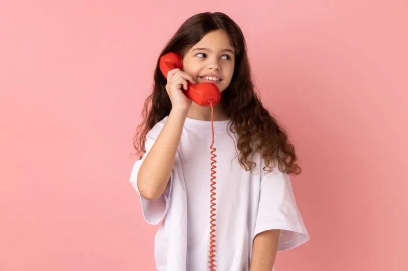 portrait of satisfied little girl wearing white t-shirt talking landline telephone holding in hand handset, looking away with toothy smile indoor studio shot isolated on pink background