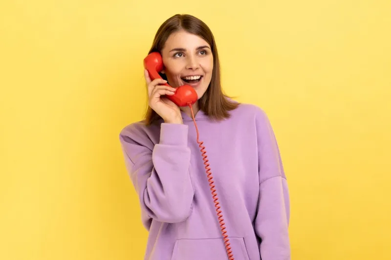 portrait of beautiful young adult woman with excited expression talking on red retro telephone, holding handset, wearing purple hoodie indoor studio shot isolated on yellow background