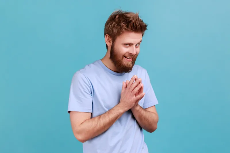 portrait of cunning bearded man schemes something keeps hands together and makes plans, sly expression looking at camera thoughtfully indoor studio shot isolated on blue background
