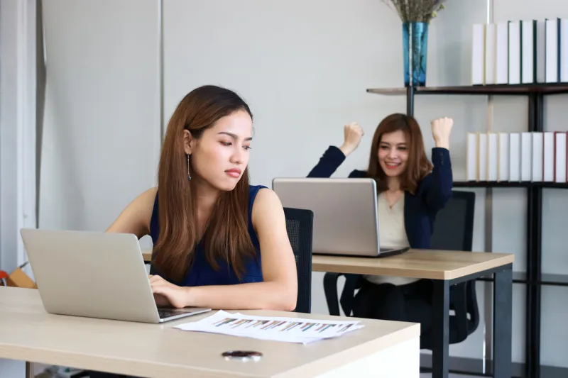 young angry envious asian business woman looking successful competitor colleague in office