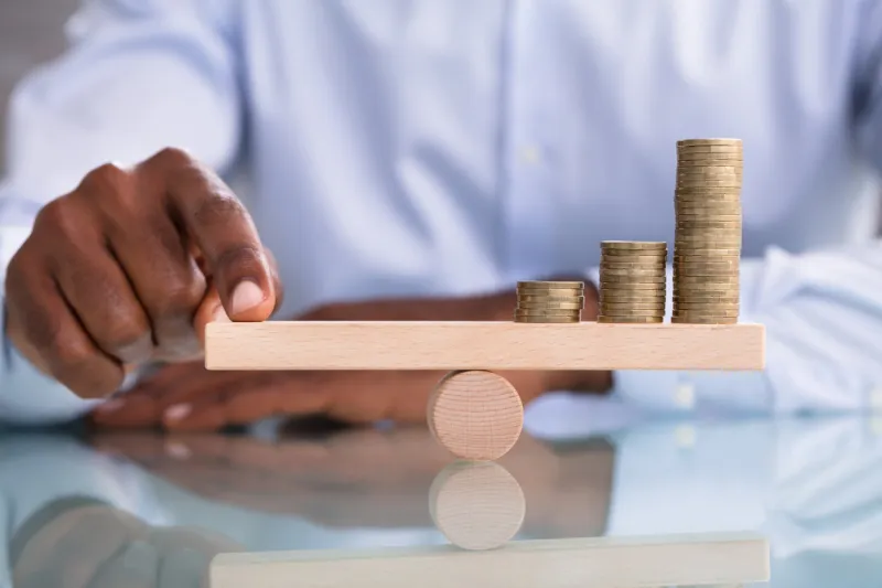 close-up of a businessman balancing the coin stack on wooden seesaw over the reflective desk